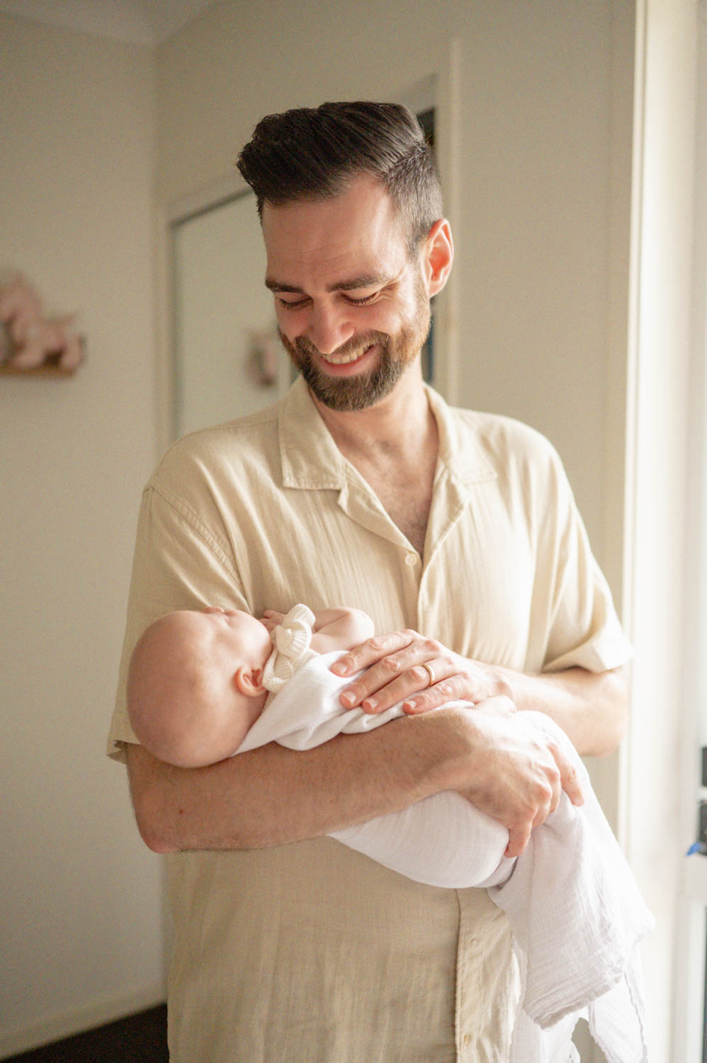 A smiling father in a cream shirt holding his newborn baby swaddled in white at home