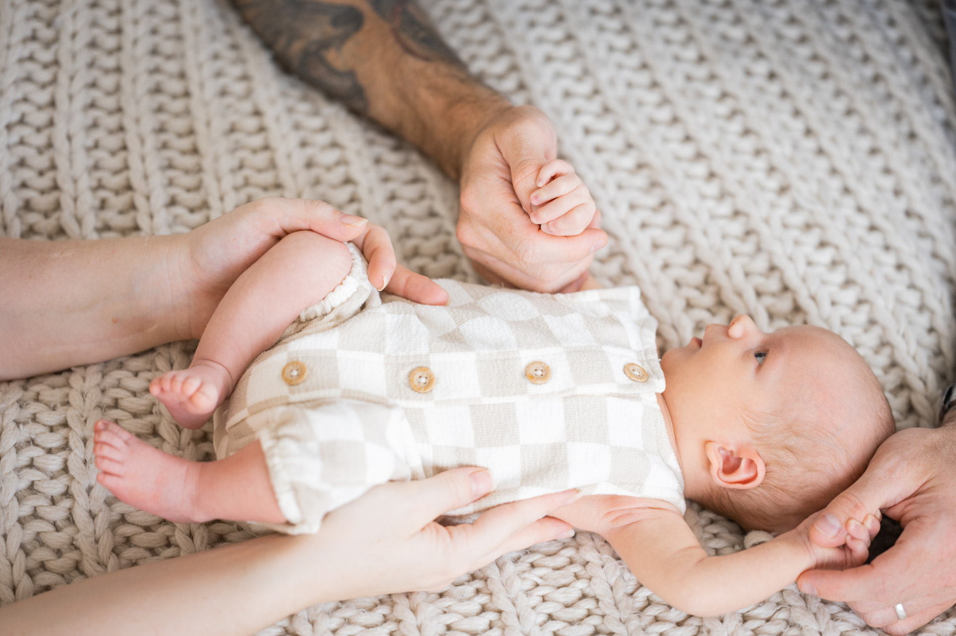 A newborn baby lying on a chunky knit blanket holding mum and dad's hands