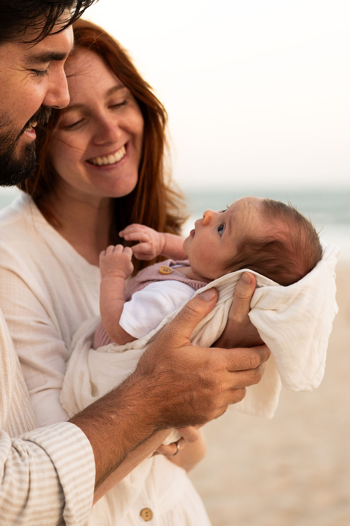 Parents smiling together on the beach as they hold their newborn baby