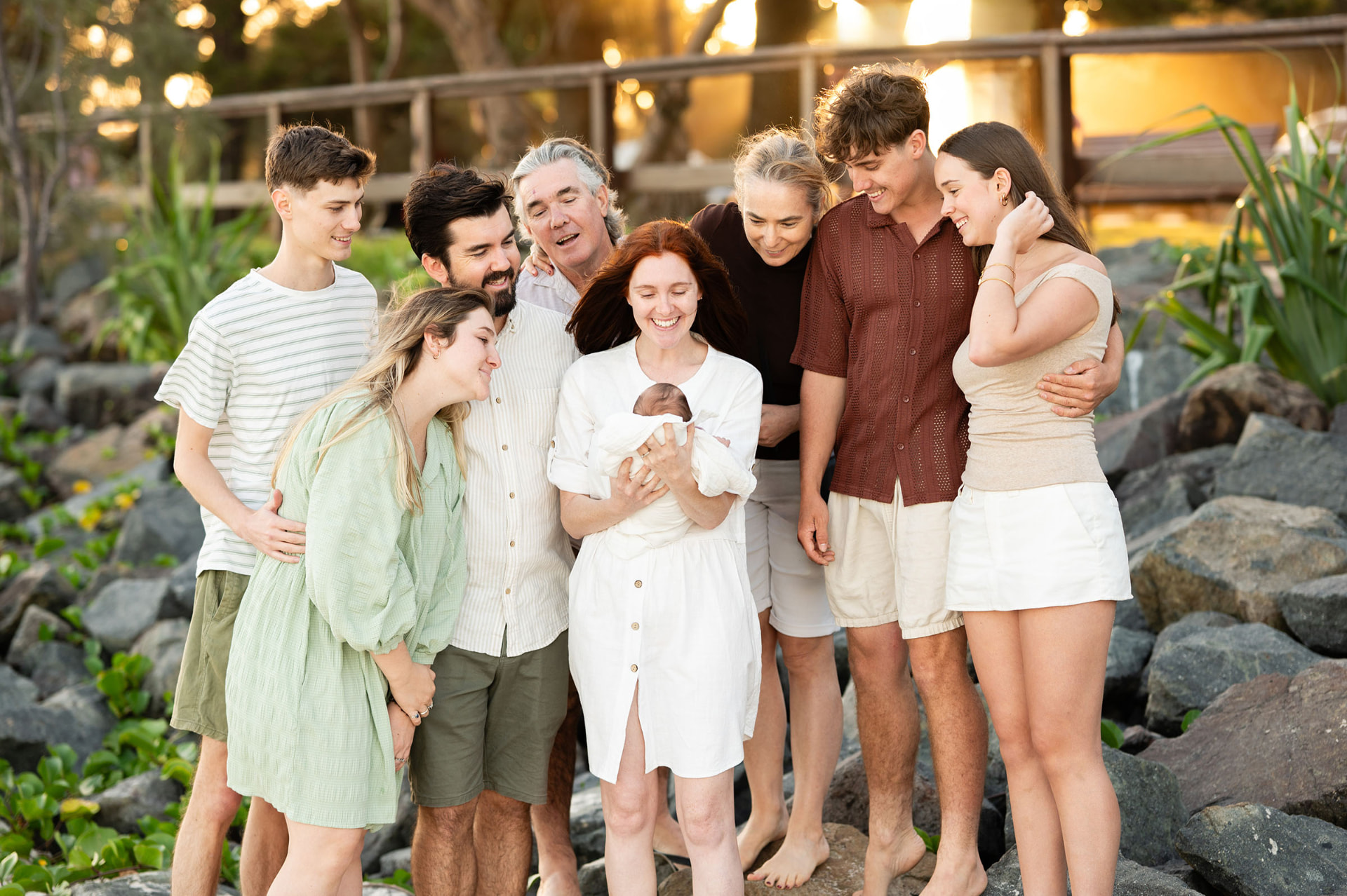An extended family gathered together on the rocks at sunset, laughing as a newborn baby is held in the middle